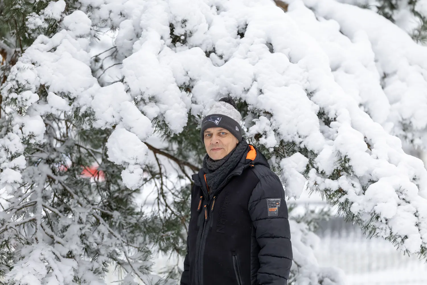 Paulchens-Hundeschule / Hunde-und Tierfotografie - Hundetraining und Fotografie - Ein Mann in einer schwarzen Winterjacke und einer Patriots-Mütze steht vor schneebedeckten Kiefernzweigen.